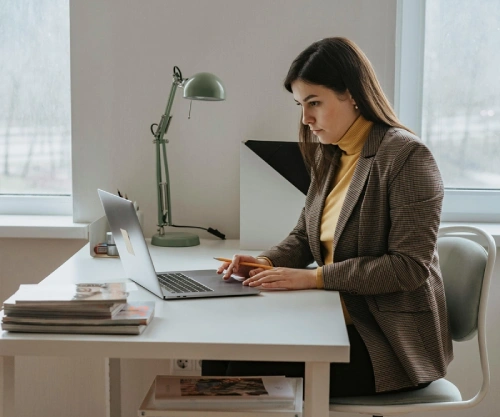 A woman is seated on a chair, focused on her assignment as she types on her laptop and holds a pencil in her right hand.