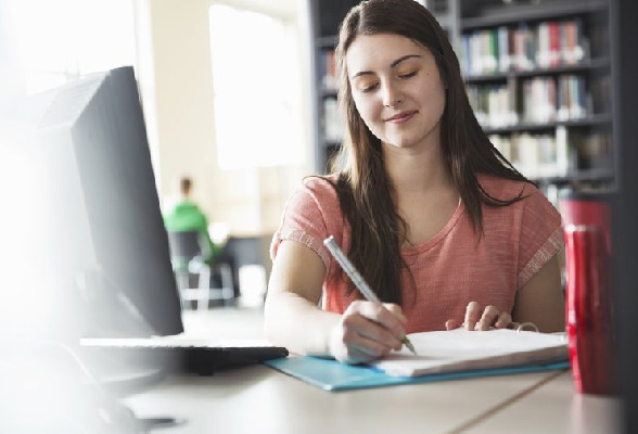 A student is seated in a classroom, working on her dissertation while jotting down notes in a notepad with a pen in her right hand.