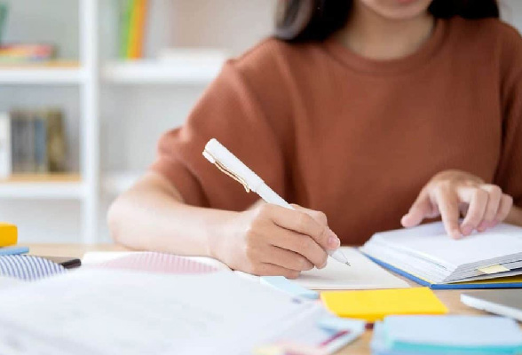 A girl is holding a pen in her right hand, working on an essay while writing notes in her notepad.